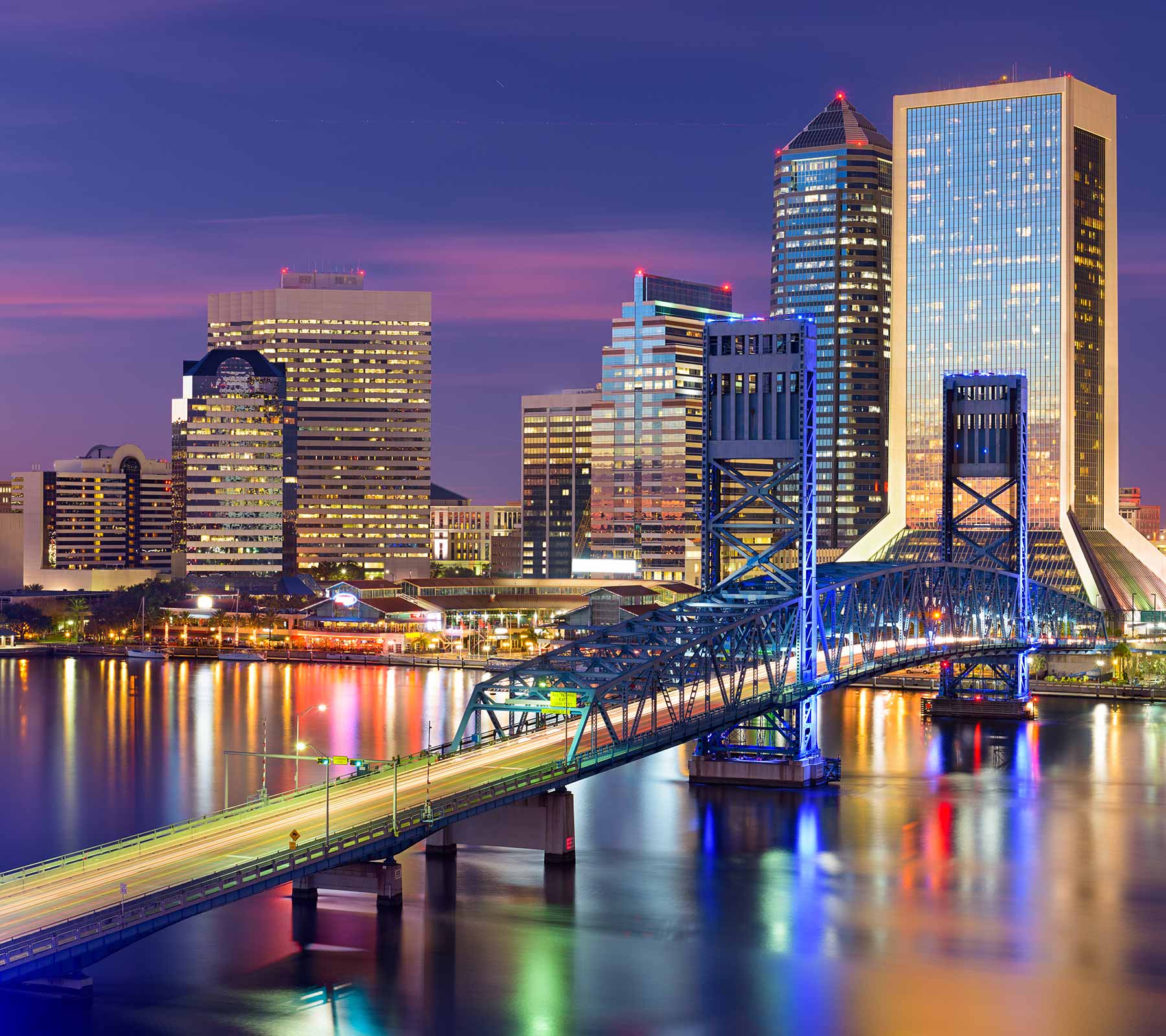City skyline at night featuring illuminated bridge and tall buildings reflected in a river.